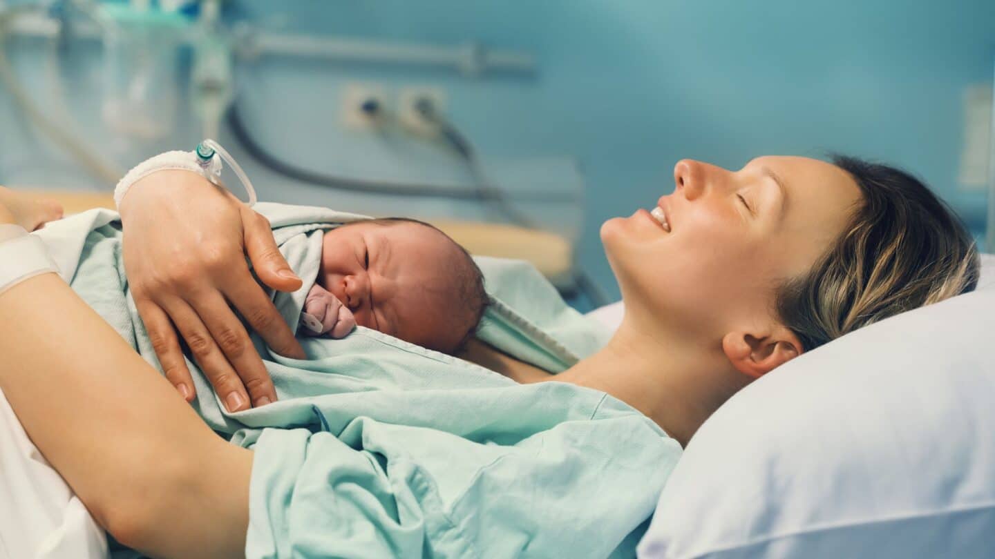 More women are using laughing gas for pain relief during labor—here’s why mother holding newborn baby in hospital bed and smiling possibly after using laughing gas for labor