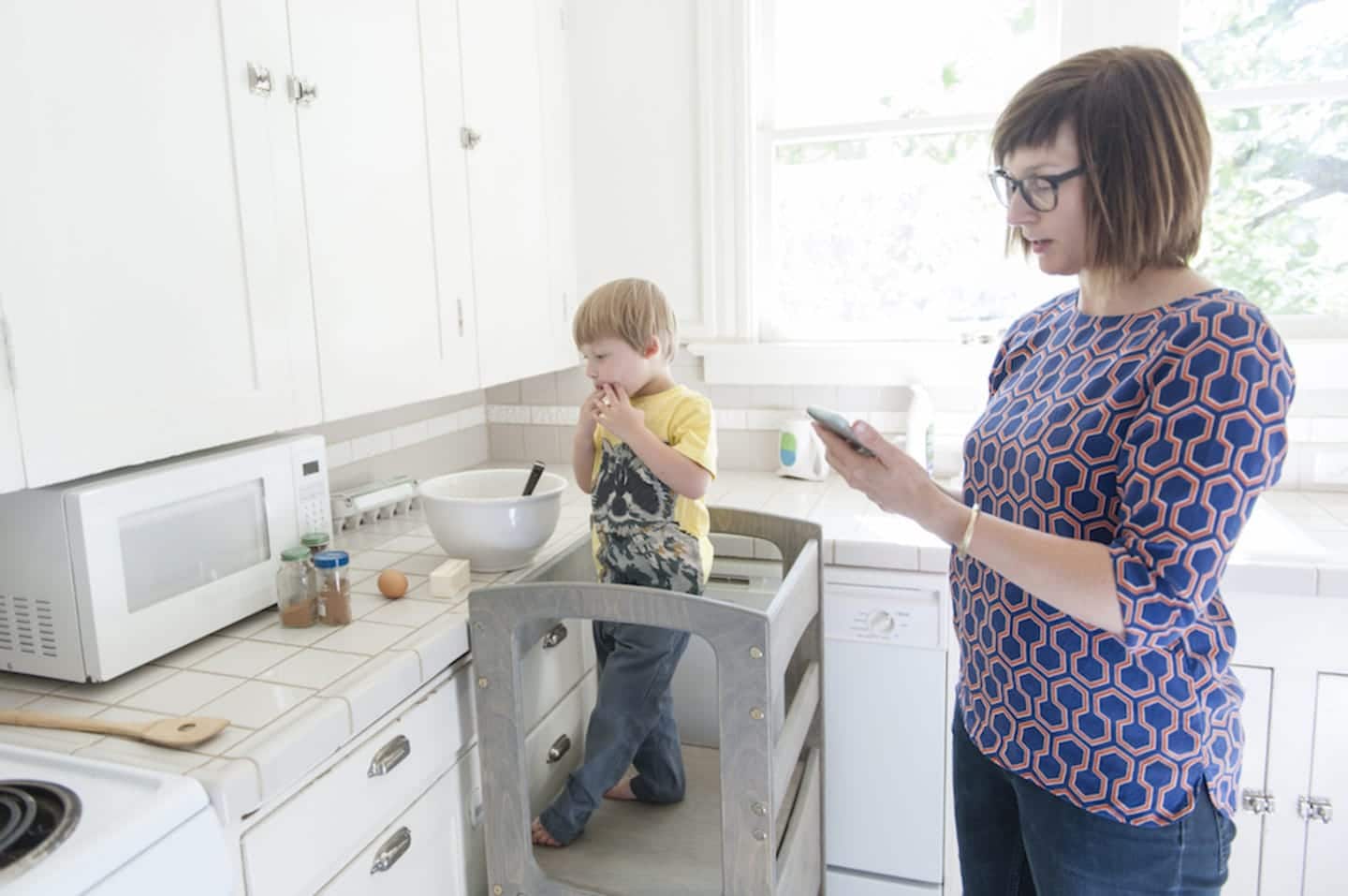 Parents are spending time with their kids—and multitasking—more than ever before woman looking at her phone and child looking at a bowl in the kitchen