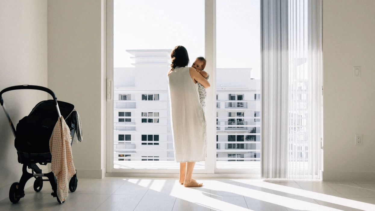 The absence of villages has made us think we need to handle motherhood alone—but we don’t mom holding baby against window doors