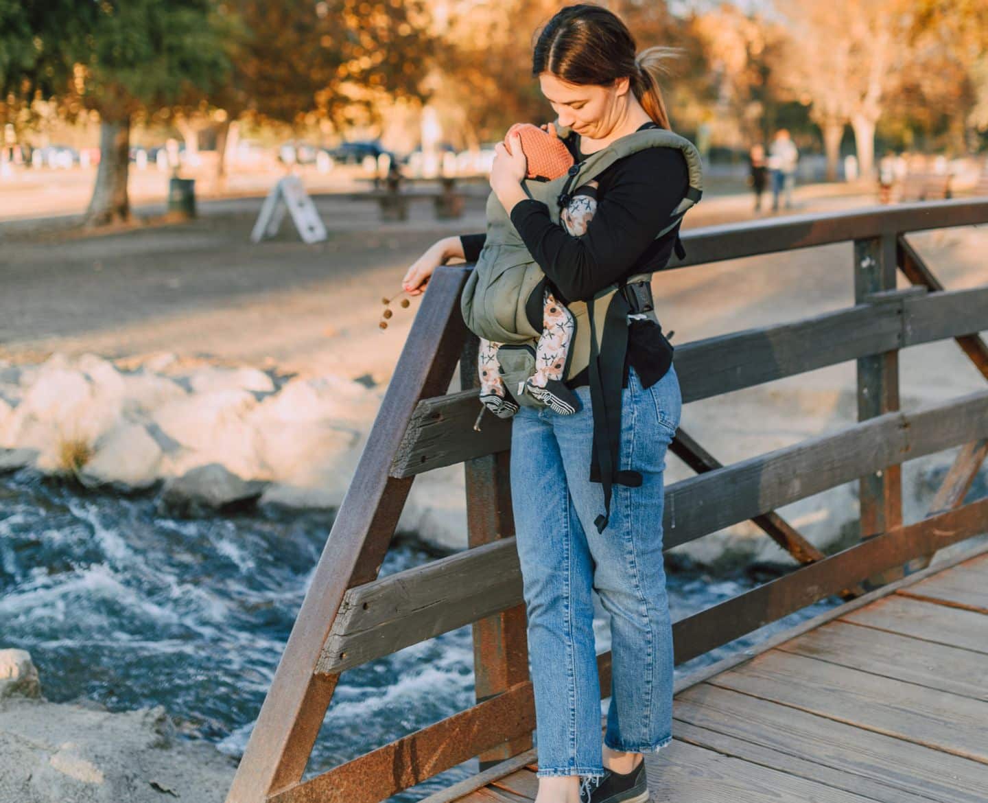 The 8 most flattering jeans for postpartum mamas woman standing on wooden bridge with her baby