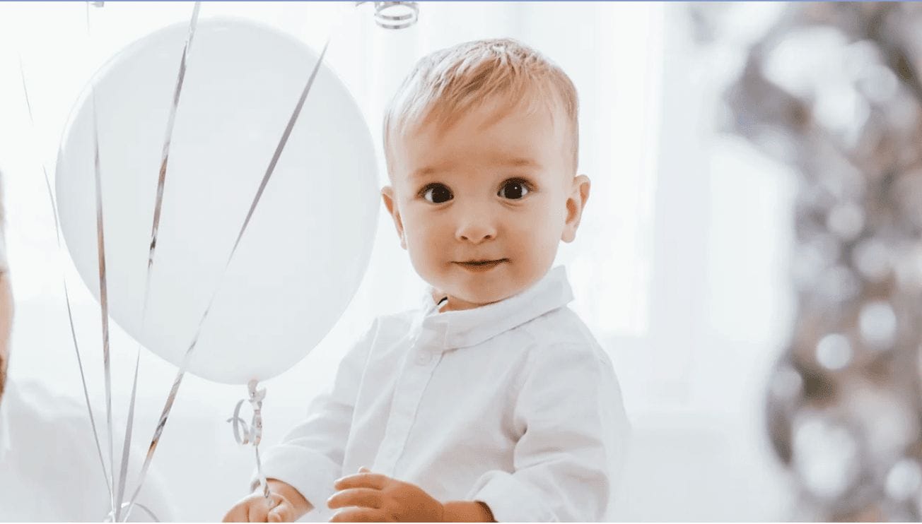 Look how far we’ve come! little boy looking at the camera with white balloons behind him