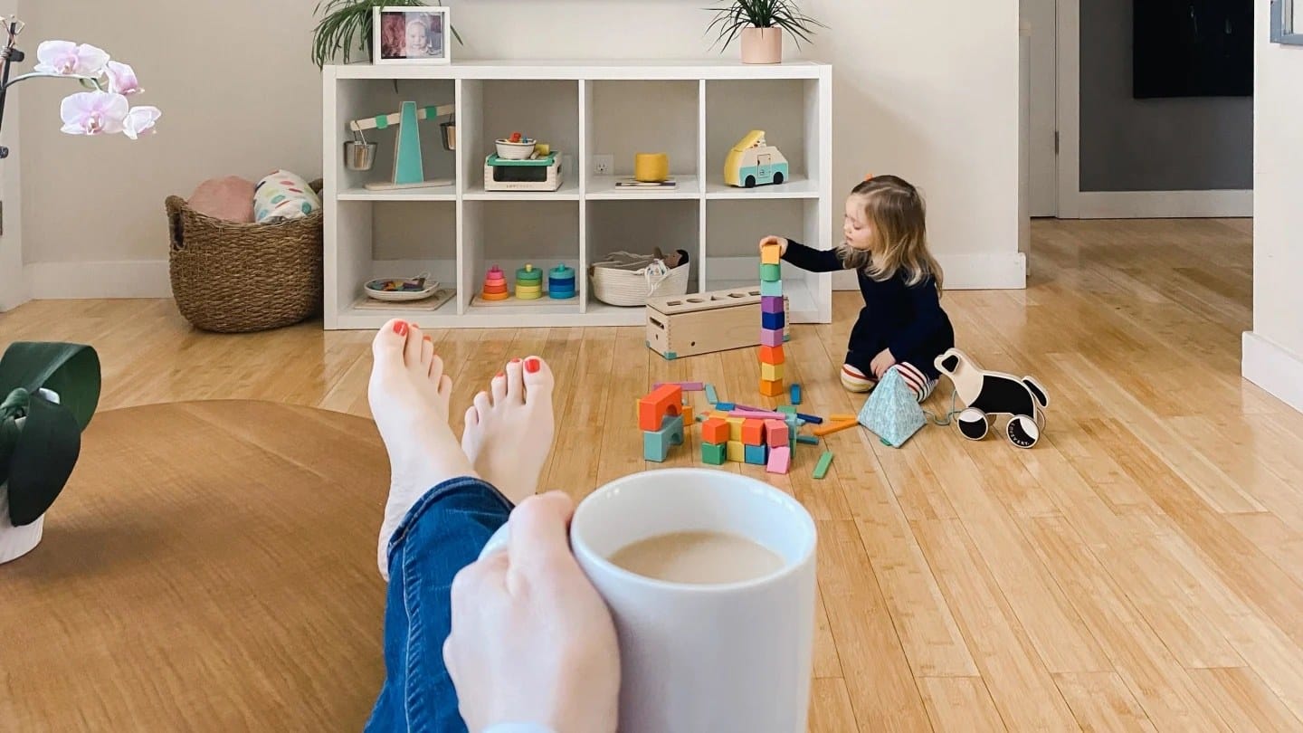 Why teaching your kids independent play is actually self-care mom holding cup of coffee with her feet up while her daughter plays with blocks in front of her
