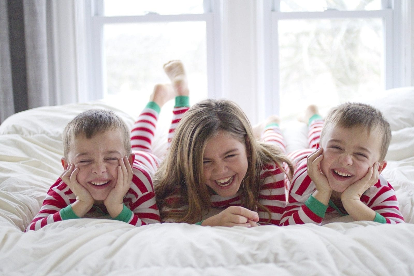 It’s true: Giving your kids fewer toys at Christmas makes them happier three kids in Christmas pajamas giggle on the bed - research shows giving kids fewer toys at Christmas contributes to happiness