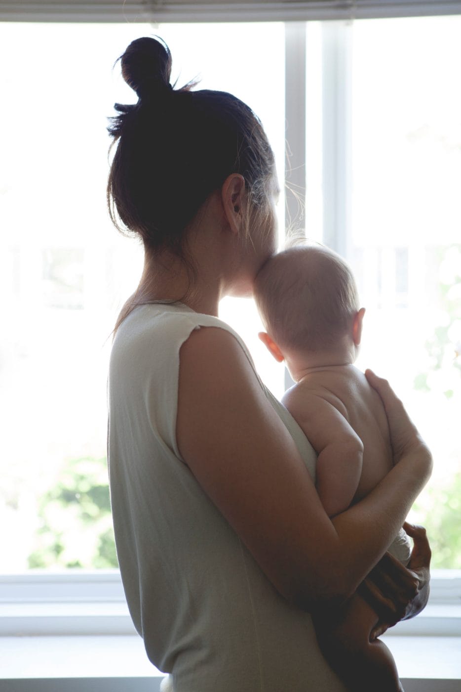 To the mamas without moms this Mother’s Day mom holding baby looking out a window as a motherless mother - mamas without moms
