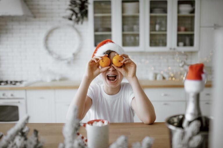 50+ holiday jokes that’ll have your kids (and you!) laughing Kid wearing a Santa hat holding oranges up to his eyes - best christmas jokes for kids