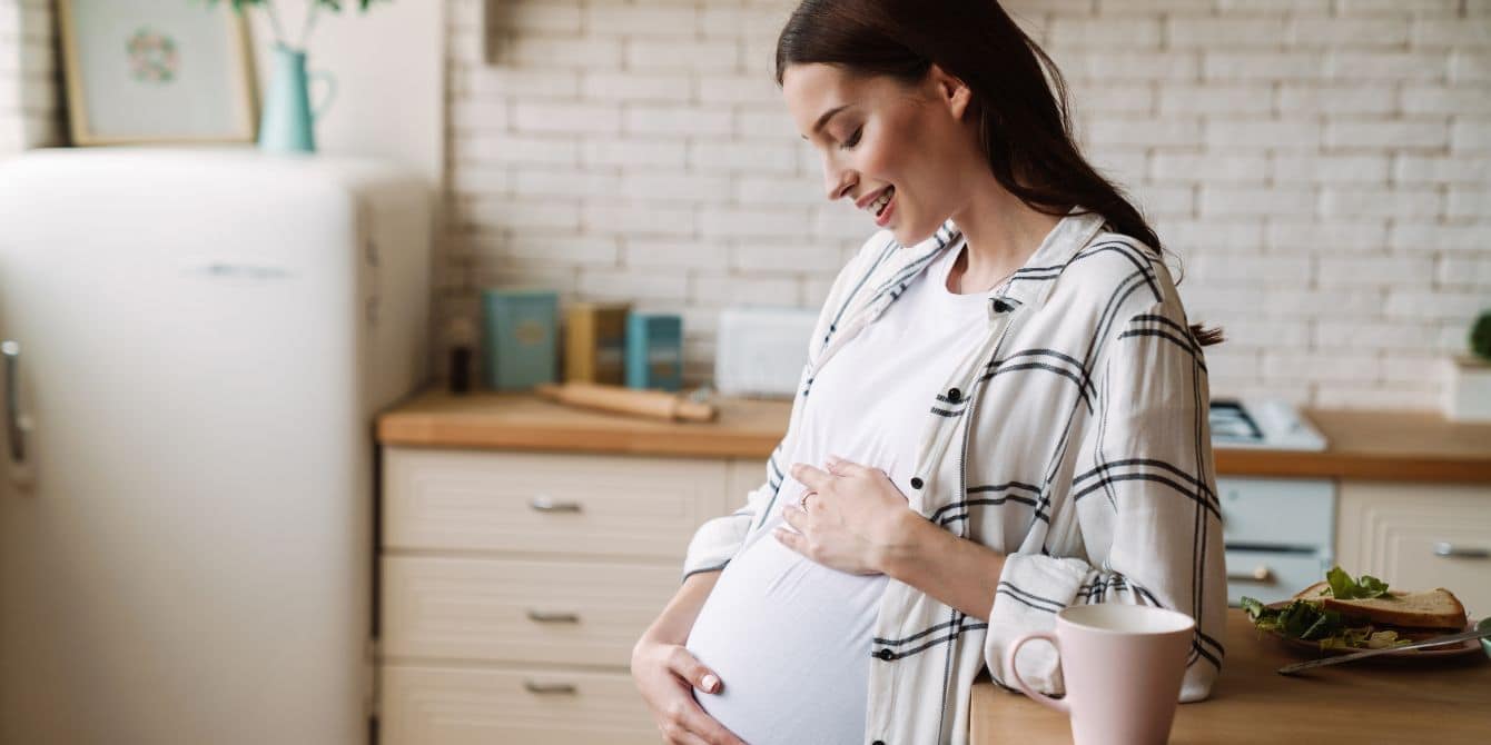 CDC warns pregnant women to avoid any deli meat or cheese unless it is reheated Pregnant woman holding belly in kitchen with sandwich on counter - listeria outbreak in deli meat and cheese