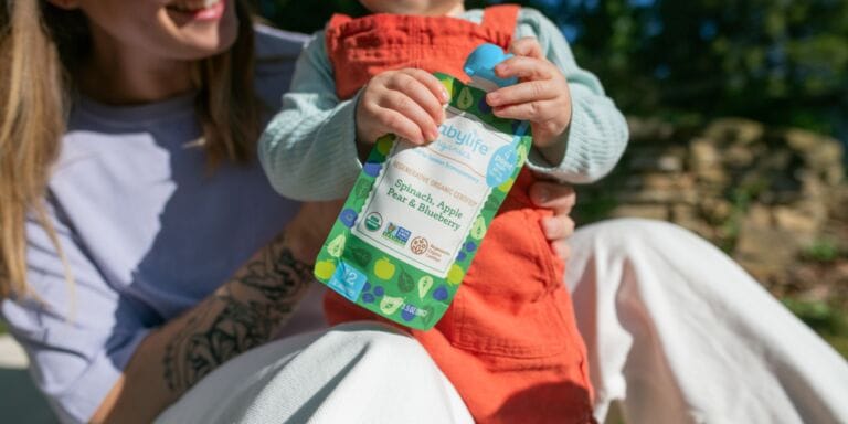This new baby food company lets you check each package for heavy metals mom holding baby who is holding a Babylife Organics baby food pouch
